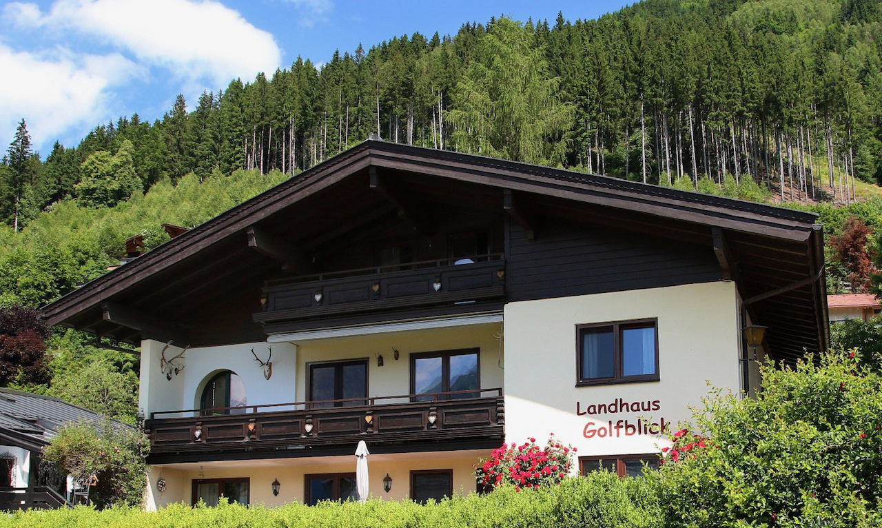 Chaletartiges Haus „Landhaus Golfblick“ vor bewaldetem Hang mit Balkonen, Hecken und blühenden Sträuchern