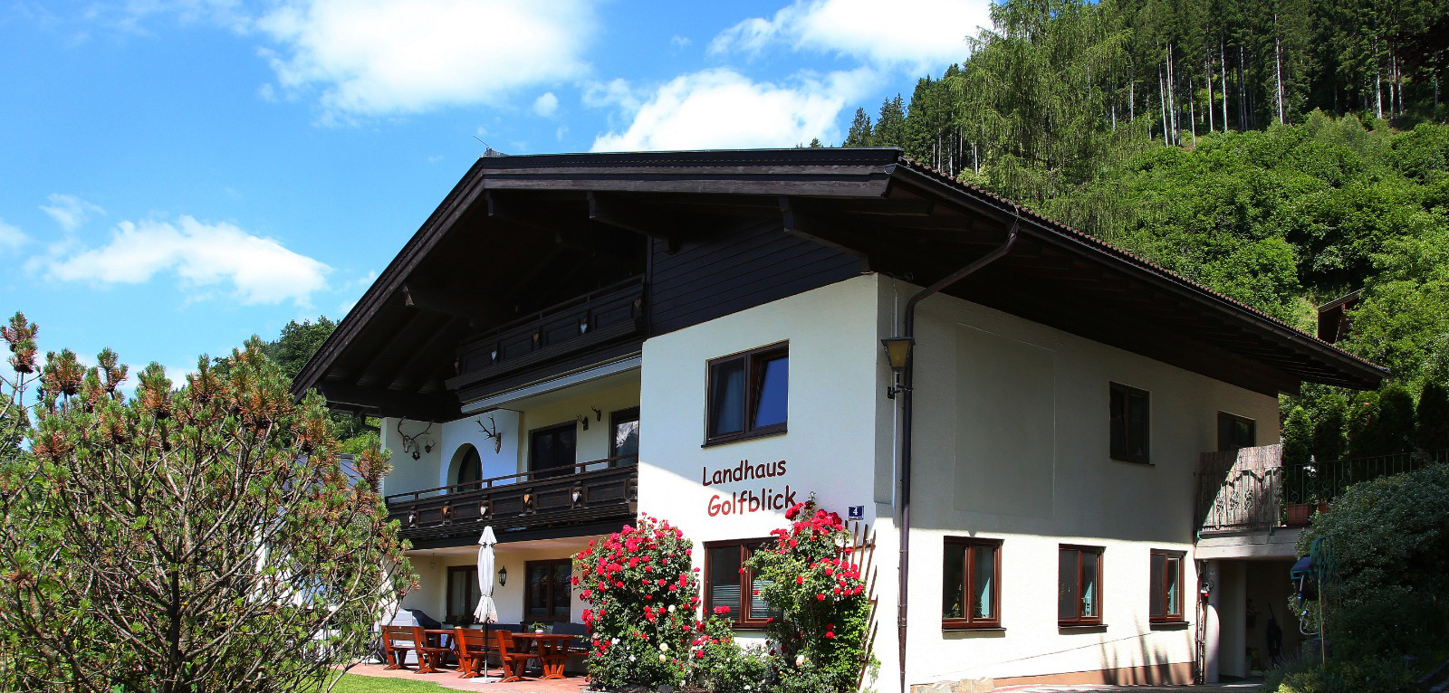 Haus mit Terrasse, Rosen und Schriftzug „Landhaus Golfblick“ vor bewaldetem Hang unter blauem Himmel